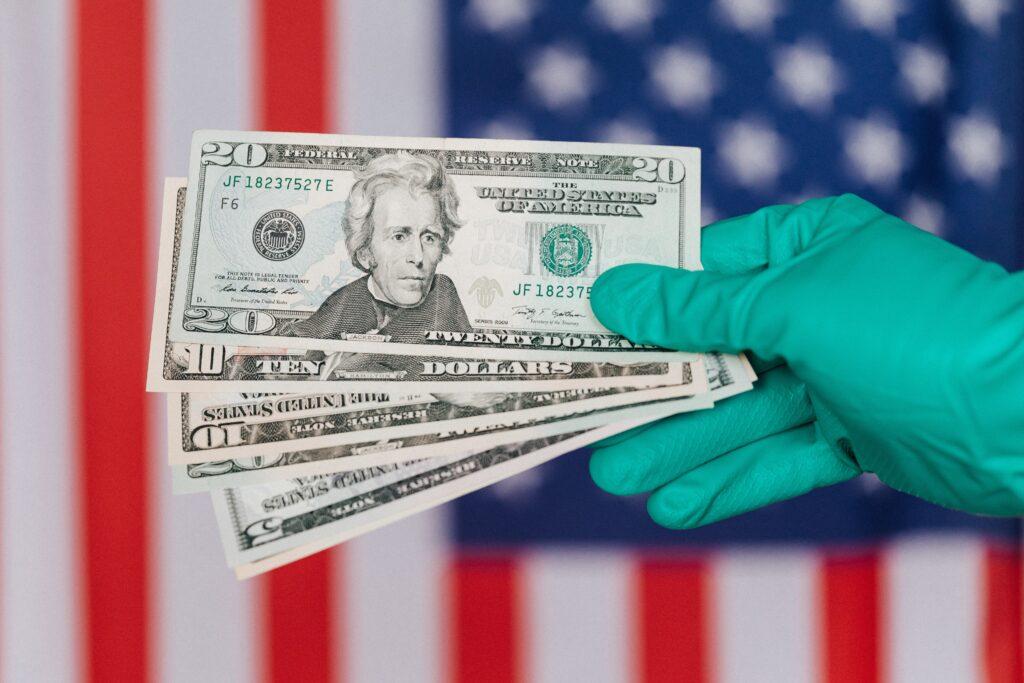 Teal colored gloved hand holding cash with an American flag background.